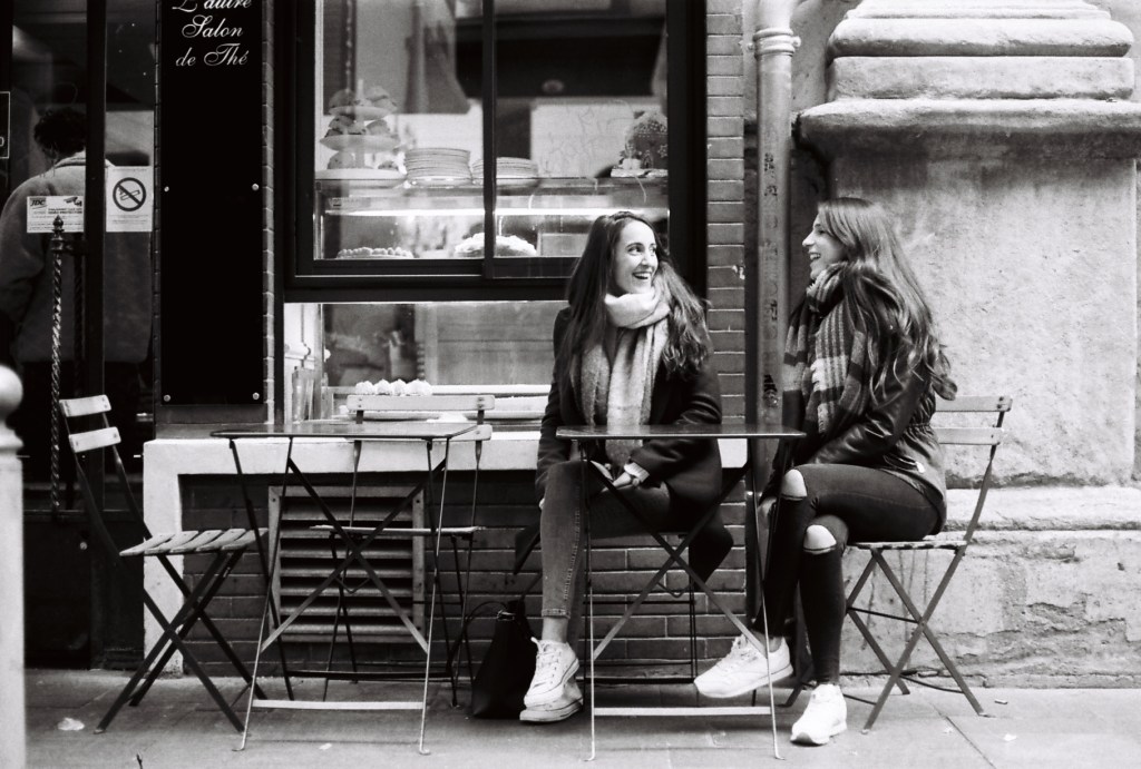 two girls sitting outside a french coffee shop laughing
